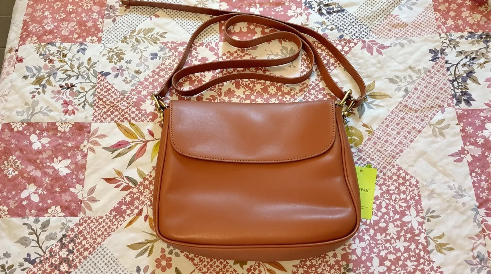 A tan leather crossbody bag lying on a patterned quilt